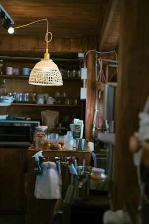 A cozy kitchen scene featuring a rustic wooden interior. Various kitchen items are placed on the counter, including a spray bottle, glass jars, a carton, and metal canisters. A mix of hanging utensils and a white cloth are visible. Overhead, a woven rattan-style light fixture is suspended, providing warm illumination.