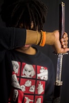 A person with a head of curly hair holds a katana vertically, with their forearm covering part of their face. They are wearing a dark sweatshirt featuring various anime-style illustrations in a grid. The person has several bracelets on their wrist, including a wooden bead bracelet and an orange one.
