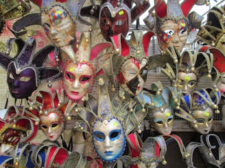 A collection of colorful Venetian masks with elaborate designs, including gold accents and intricate patterns, displayed on a mesh background. Each mask features unique embellishments such as bells and fabrics, creating a vibrant and festive appearance.