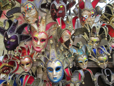 A collection of colorful Venetian masks with elaborate designs, including gold accents and intricate patterns, displayed on a mesh background. Each mask features unique embellishments such as bells and fabrics, creating a vibrant and festive appearance.