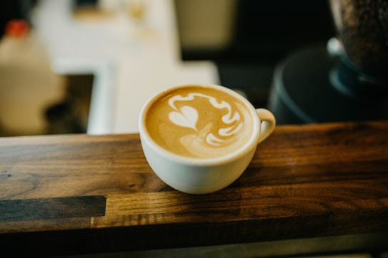 A cup of latte with intricate latte art on its surface, resting on a wooden countertop. The background is slightly blurred, suggesting a cozy coffee shop ambiance.