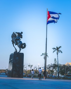 A large equestrian statue on a high pedestal stands prominently against a clear blue sky. Nearby, a tall flagpole displays a red, white, and blue flag. People walk around the open plaza, bordered by trees and distant vehicles.