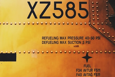 Close-up view of an aircraft or industrial vehicle surface with rivets and textual markings. The markings include specifications regarding fuel and pressure limits, presented in black text on a yellow background. The area is coated with yellow paint and features a prominent alphanumeric designation XZ585.
