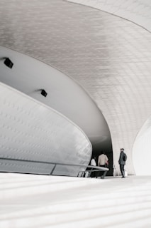 A modern architectural space featuring curved, sleek white surfaces and a geometric ceiling. Several people can be seen walking through the structure, adding a sense of scale. The area is illuminated by natural light, creating soft shadows and highlights.