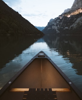 A canoe is positioned in the foreground, pointed towards a serene, reflective body of water surrounded by lush green forests and towering rocky mountains. The calm water mirrors the landscape, creating a symmetrical and tranquil scene.