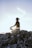 A person is sitting on a rocky surface outdoors, practicing meditation with sunlight shining through their arms. The sky is clear and the sun is low, creating a serene and peaceful atmosphere.