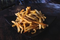 A pile of golden-brown French fries is placed on a black plate. The fries appear crispy and are resting on a dark, textured surface, likely a wooden table.
