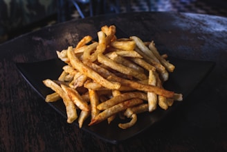 A pile of golden-brown French fries is placed on a black plate. The fries appear crispy and are resting on a dark, textured surface, likely a wooden table.