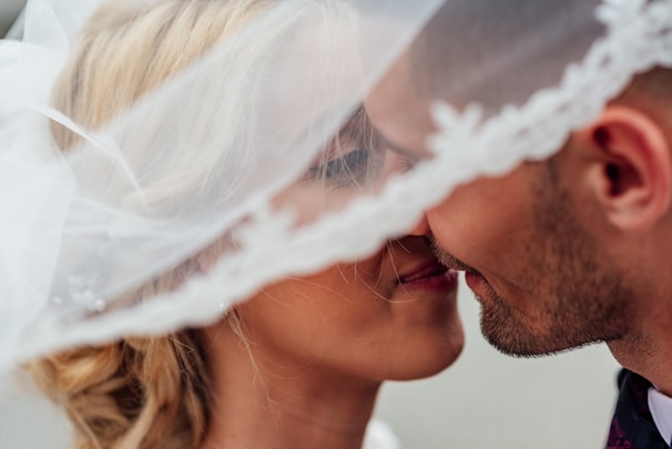A close-up shot of a couple about to kiss, partially covered by a white veil. The woman has blonde hair styled in an elegant updo, and the man has short dark hair with a beard. The soft focus and intimate setting convey a romantic moment.