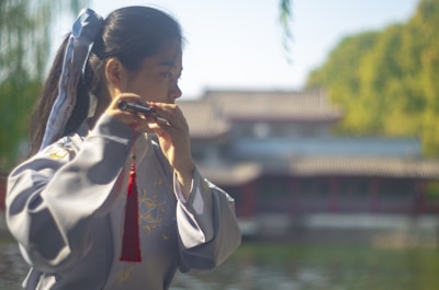 A person in traditional clothing plays a flute in an outdoor setting. Their hair is tied with a ribbon, and they are standing near a body of water with trees and a traditional building visible in the background.