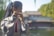A person in traditional clothing plays a flute in an outdoor setting. Their hair is tied with a ribbon, and they are standing near a body of water with trees and a traditional building visible in the background.