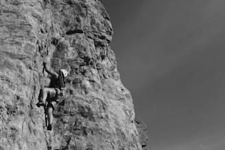 A person wearing a helmet and climbing gear is ascending a steep rock face, showcasing a challenging rock climbing activity. The rock is rugged and tall, and the climber is focused on their ascent, gripping onto the rock surface. The sky in the background is clear, providing a stark contrast to the detailed texture of the rock.