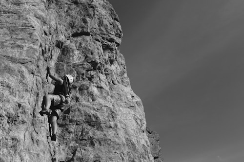 A person wearing a helmet and climbing gear is ascending a steep rock face, showcasing a challenging rock climbing activity. The rock is rugged and tall, and the climber is focused on their ascent, gripping onto the rock surface. The sky in the background is clear, providing a stark contrast to the detailed texture of the rock.