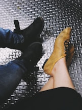 Two pairs of shoes are visible on a textured metallic floor. One pair is black sneakers paired with blue jeans, and the other is yellow perforated shoes worn by someone with bare legs.
