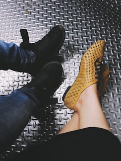 Two pairs of shoes are visible on a textured metallic floor. One pair is black sneakers paired with blue jeans, and the other is yellow perforated shoes worn by someone with bare legs.