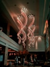A luxurious interior space featuring an extravagant chandelier with swirling, illuminated designs hanging from a high ceiling. The area is warmly lit, with elegant furnishings and large vases containing decorative plants. Soft lighting and modern decor create a sophisticated atmosphere.