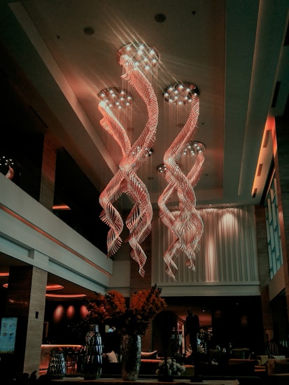 A luxurious interior space featuring an extravagant chandelier with swirling, illuminated designs hanging from a high ceiling. The area is warmly lit, with elegant furnishings and large vases containing decorative plants. Soft lighting and modern decor create a sophisticated atmosphere.