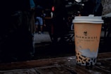 A disposable coffee cup with a logo and text, placed on a wet wooden surface. The background features a group of people casually dressed, with some wearing shorts. The setting appears to be outdoors, likely in a public space or market.