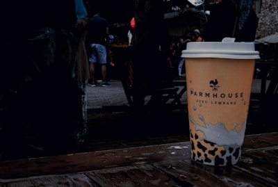 A disposable coffee cup with a logo and text, placed on a wet wooden surface. The background features a group of people casually dressed, with some wearing shorts. The setting appears to be outdoors, likely in a public space or market.