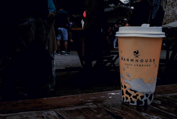 A disposable coffee cup with a logo and text, placed on a wet wooden surface. The background features a group of people casually dressed, with some wearing shorts. The setting appears to be outdoors, likely in a public space or market.