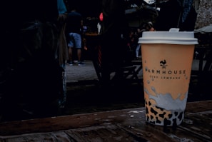 A disposable coffee cup with a logo and text, placed on a wet wooden surface. The background features a group of people casually dressed, with some wearing shorts. The setting appears to be outdoors, likely in a public space or market.