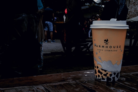 A disposable coffee cup with a logo and text, placed on a wet wooden surface. The background features a group of people casually dressed, with some wearing shorts. The setting appears to be outdoors, likely in a public space or market.