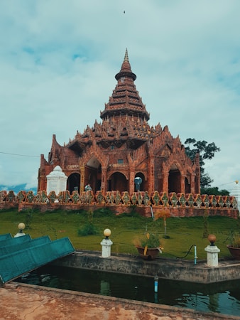 A richly decorated temple with intricate carvings and a tiered spire stands against a partly cloudy sky. The temple is surrounded by a lush green lawn and a low stone wall adorned with decorative elements. In front of the temple, there's a small water feature with a stone edge and some aquatic plants.