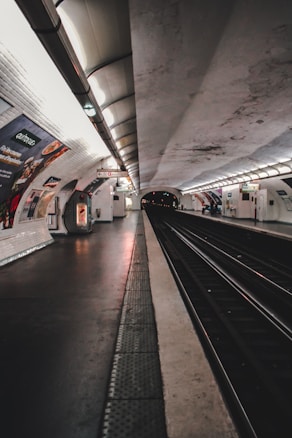 A subway platform with tiled walls and ceiling, featuring advertisements on the walls. The platform is empty, with tracks running down the center. Overhead lighting creates a dim ambiance, and there are signs and a small vending machine visible.