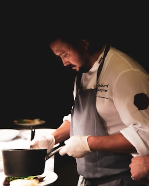 A chef wearing an apron and gloves is focused on preparing a dish in a dimly lit kitchen. The chef holds a pot over a plate, carefully arranging the food. The atmosphere is intimate and concentrated, emphasizing the art and skill of culinary creation.