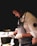 A chef wearing an apron and gloves is focused on preparing a dish in a dimly lit kitchen. The chef holds a pot over a plate, carefully arranging the food. The atmosphere is intimate and concentrated, emphasizing the art and skill of culinary creation.
