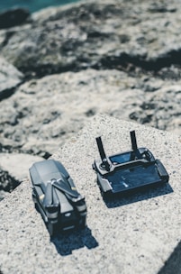 A compact drone and its controller are placed on a textured stone surface. The background consists of blurred rock formations, hinting at an outdoor setting.