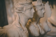 Three stone busts with classical features are lined up closely together. The figures are chiseled with precise detail, capturing the contours of their faces and hair. The warm lighting creates a soft and muted atmosphere, enhancing the aged appearance of the sculptures.