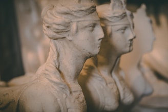 Three stone busts with classical features are lined up closely together. The figures are chiseled with precise detail, capturing the contours of their faces and hair. The warm lighting creates a soft and muted atmosphere, enhancing the aged appearance of the sculptures.