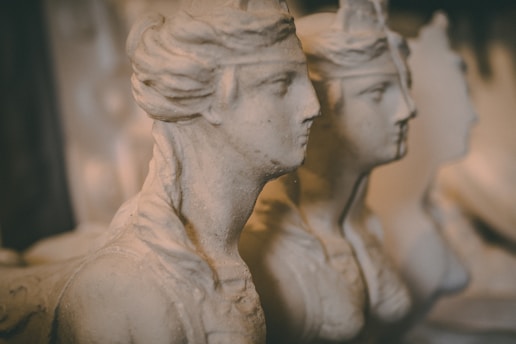 Three stone busts with classical features are lined up closely together. The figures are chiseled with precise detail, capturing the contours of their faces and hair. The warm lighting creates a soft and muted atmosphere, enhancing the aged appearance of the sculptures.