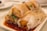 A close-up view of rice noodle rolls filled with fried dough sticks, topped with sesame seeds, and garnished with chopped green onions. The rolls are drizzled with soy sauce, creating a glossy texture. The dish is served on a rectangular white plate.