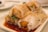A close-up view of rice noodle rolls filled with fried dough sticks, topped with sesame seeds, and garnished with chopped green onions. The rolls are drizzled with soy sauce, creating a glossy texture. The dish is served on a rectangular white plate.