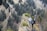 A group of people are hiking along a narrow, rocky path on a mountainside covered with dense evergreen trees. The landscape is rugged, with steep slopes and dramatic cliffs, creating a sense of adventure and wilderness.