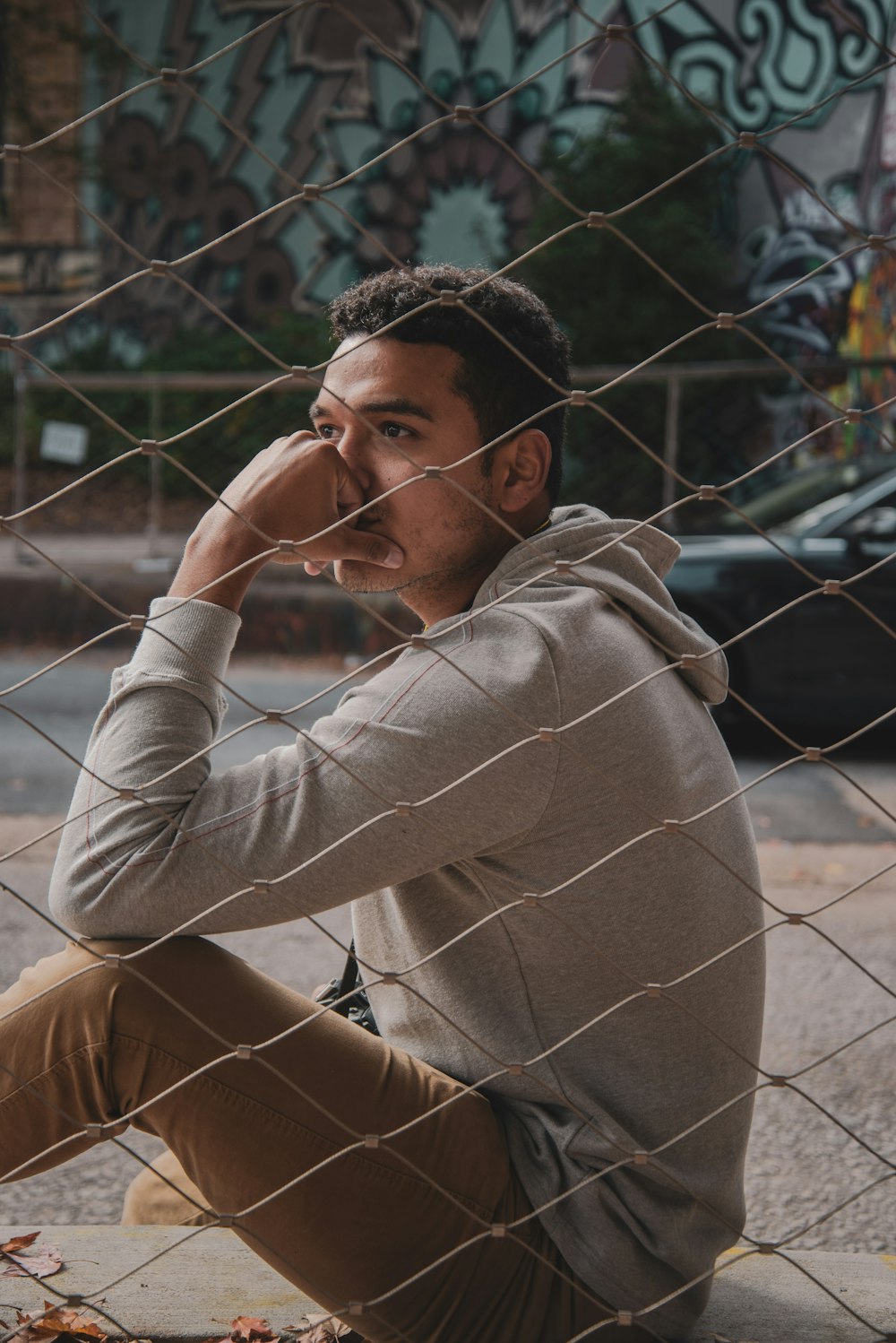 man sitting beside fence with hand on face outdoors during daytime