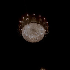A large, ornate crystal chandelier emits a warm glow against a dark background. The chandelier is detailed with numerous sparkling crystals and several candle-like light bulbs around it, creating an elegant and luxurious ambiance.