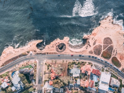 Aerial view of a coastal landscape featuring a road with cars running parallel to the shoreline. On one side of the road, there are several residential houses with red and white rooftops, surrounded by greenery. The coast is rocky with distinct wave patterns in the water, and a sandy area with visible pathways.