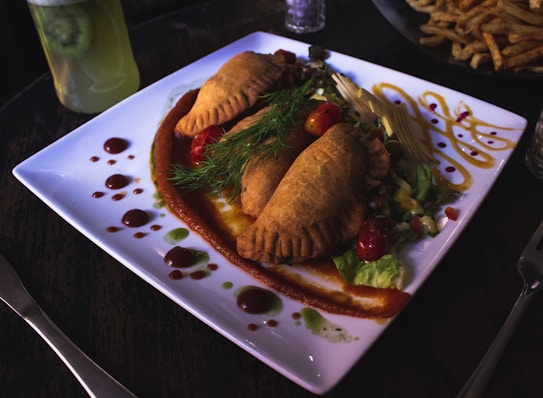 A white square plate features a neatly arranged dish of pastries filled with a savory stuffing, garnished with sprigs of dill. The plate also includes leafy greens and sliced vegetables on the side, with drizzled sauces in green and red patterns adding a decorative touch. A glass of kiwi drink is visible in the background, along with a bowl of fries.