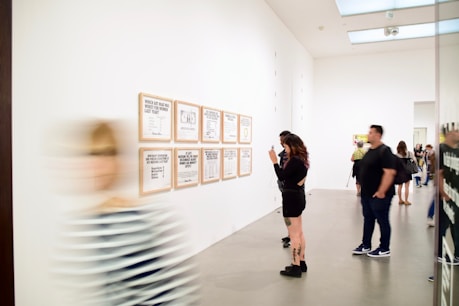 A modern art gallery with people viewing framed text-based artworks on a white wall. One person is taking a photo of the artworks while others are looking at them. A blurred figure is walking past in the foreground.