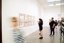 A modern art gallery with people viewing framed text-based artworks on a white wall. One person is taking a photo of the artworks while others are looking at them. A blurred figure is walking past in the foreground.