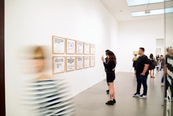 A modern art gallery with people viewing framed text-based artworks on a white wall. One person is taking a photo of the artworks while others are looking at them. A blurred figure is walking past in the foreground.