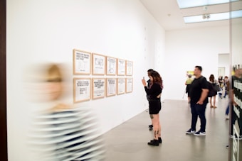 A modern art gallery with people viewing framed text-based artworks on a white wall. One person is taking a photo of the artworks while others are looking at them. A blurred figure is walking past in the foreground.