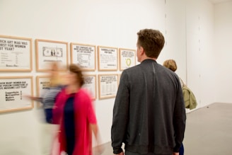 Several people are observing framed informational posters on a wall in an art gallery. The posters contain text and graphics related to women in art. The scene is indoors with a simple, clean setting. A few artworks have text addressing gender issues in the art world.