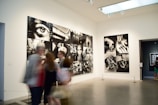 A gallery with modern art photographs displayed on white walls, featuring large black and white images with various abstract and surreal elements. Several people are walking past, slightly blurred, indicating motion.