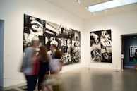 A gallery with modern art photographs displayed on white walls, featuring large black and white images with various abstract and surreal elements. Several people are walking past, slightly blurred, indicating motion.