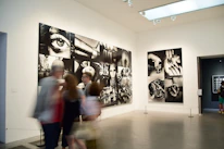 A gallery with modern art photographs displayed on white walls, featuring large black and white images with various abstract and surreal elements. Several people are walking past, slightly blurred, indicating motion.