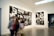 A gallery with modern art photographs displayed on white walls, featuring large black and white images with various abstract and surreal elements. Several people are walking past, slightly blurred, indicating motion.